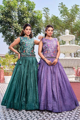 Two women in green and purple traditional dresses standing outdoors with a fountain in the background.