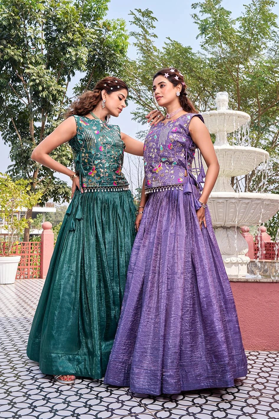 Two women in green and purple traditional dresses standing outdoors with a fountain in the background.