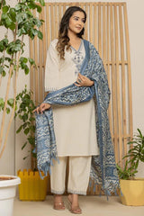 Woman in a cream outfit with a blue patterned scarf standing indoors with plants around.