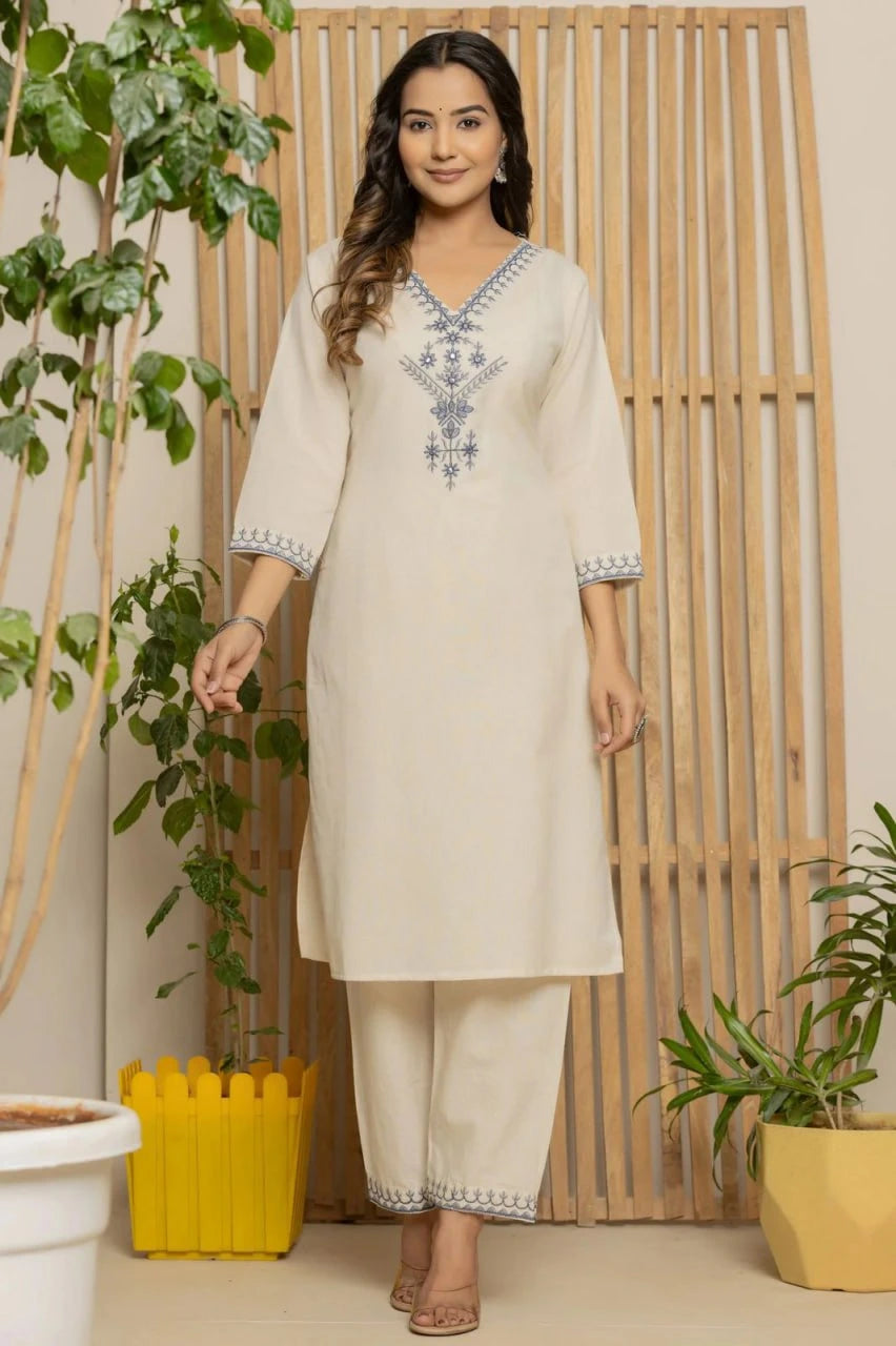 Woman wearing a cream kurta with blue embroidery standing in a room with plants and wooden decor.