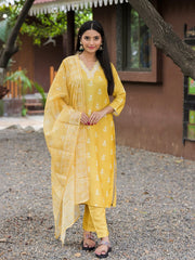 Woman in a yellow traditional outfit standing outdoors with a brown building and greenery in the background.