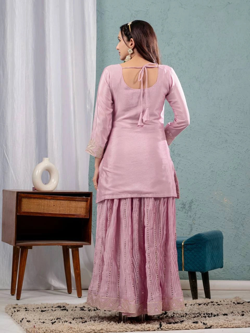 Woman wearing a pink traditional outfit standing in a room with a side table and chair.