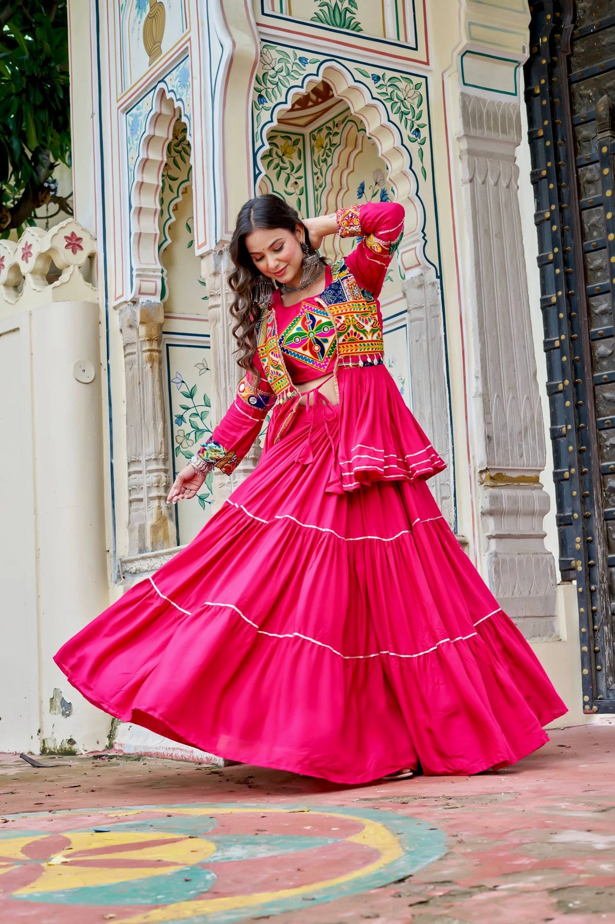 Woman in a bright pink traditional outfit with colorful embroidery standing in front of an ornate architectural background.