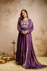 Woman in a purple traditional outfit standing against a textured beige wall.