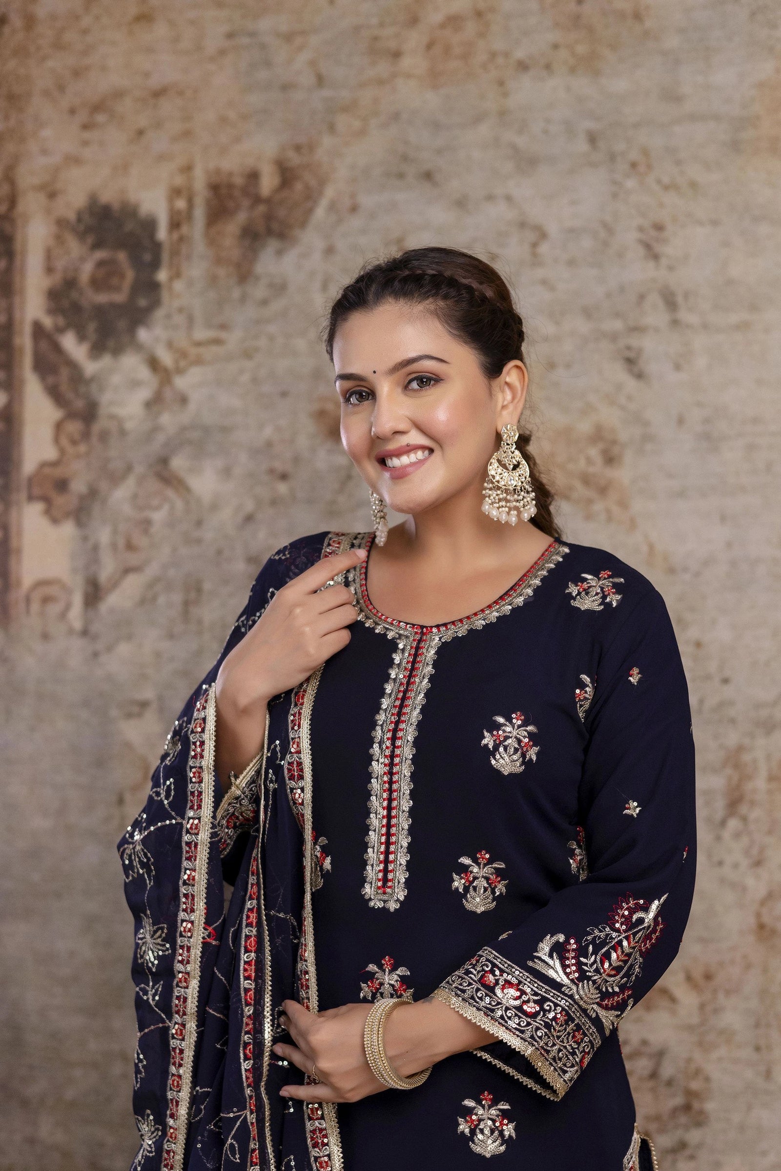 Woman wearing a navy blue embroidered traditional outfit against a textured wall.