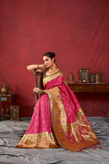 A woman models a luxurious Hot Pink Soft Banarasi Silk Saree. The saree features dense, all-over tone-on-tone Zari weaving. The wide border and pallu showcase heavy gold Zari work with vibrant green and yellow accents. She wears a contrasting dark (black/brown) sleeveless blouse and a heavy gold Kundan choker necklace, posing indoors.