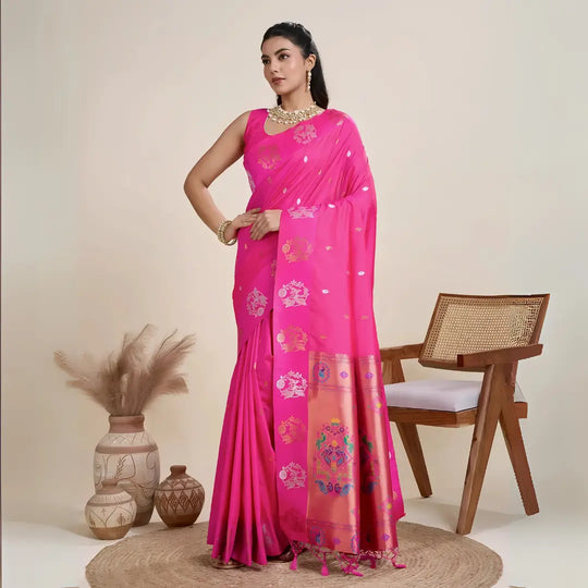 A woman models a luxurious Rani Pink Soft Silk Saree. The saree features scattered metallic motifs and a wide pallu in contrasting copper/rose gold, richly woven with multi-colored peacock and floral designs. She wears a matching pink sleeveless blouse and heavy gold Kundan jewelry, posing indoors.