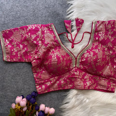 Pink brocade blouse featuring intricate gold floral zari work and decorative trim along the neckline and sleeves, displayed flat on a grey surface with a white faux fur accent and a bunch of artificial pink and purple flowers for an elegant, festive ethnic look.