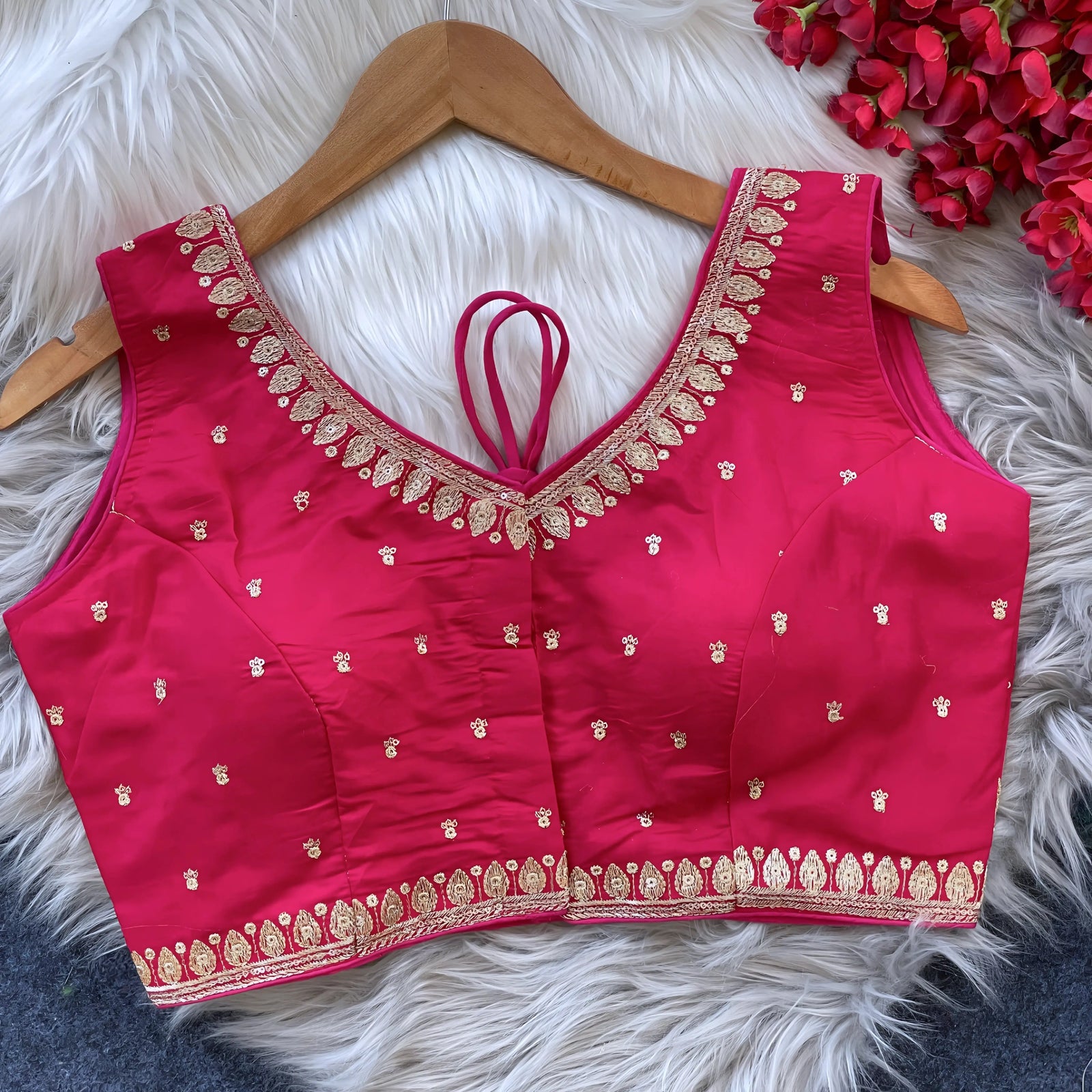 Pink padded blouse with intricate gold embroidery along the neckline and hem, featuring small gold motifs, displayed on a wooden hanger over a white fur surface with red artificial flowers in the background.