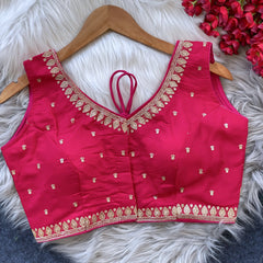 Pink padded blouse with intricate gold embroidery along the neckline and hem, featuring small gold motifs, displayed on a wooden hanger over a white fur surface with red artificial flowers in the background.