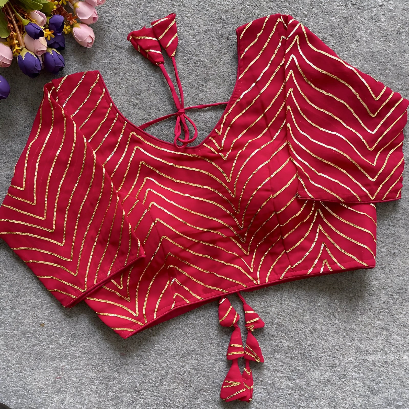 Pink blouse featuring striking gold gota patti zigzag embroidery, three-quarter sleeves, and a stylish back tie-up with matching triangular tassels. Displayed flat on a textured grey surface beside fresh pink and purple flowers, emphasizing its festive and elegant ethnic appeal.
