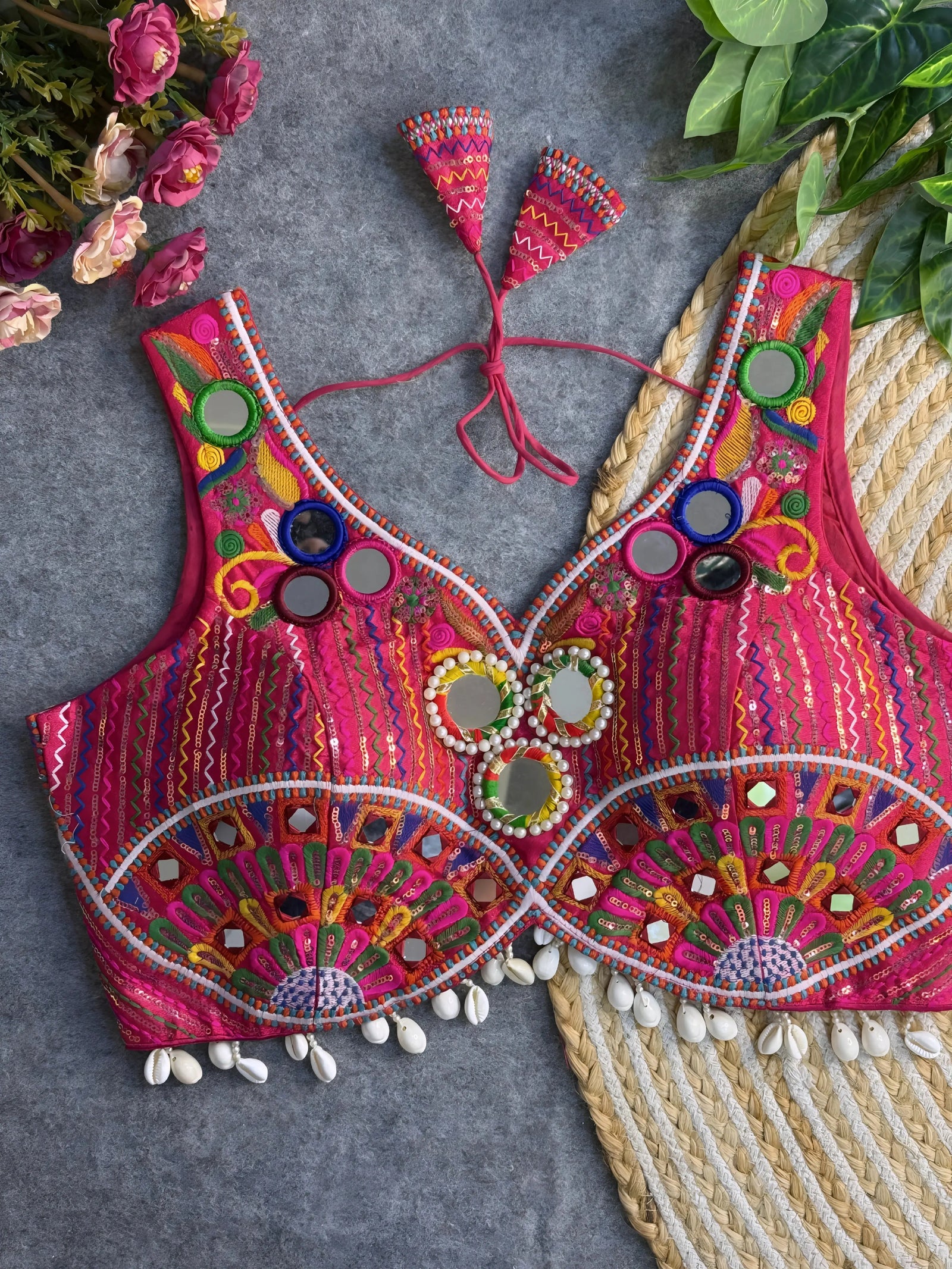Traditional pink embroidered blouse featuring vibrant multicolor mirror work, intricate thread embroidery in geometric and floral patterns, and shell tassel detailing along the hem, displayed on a flat surface with floral and leafy decor.