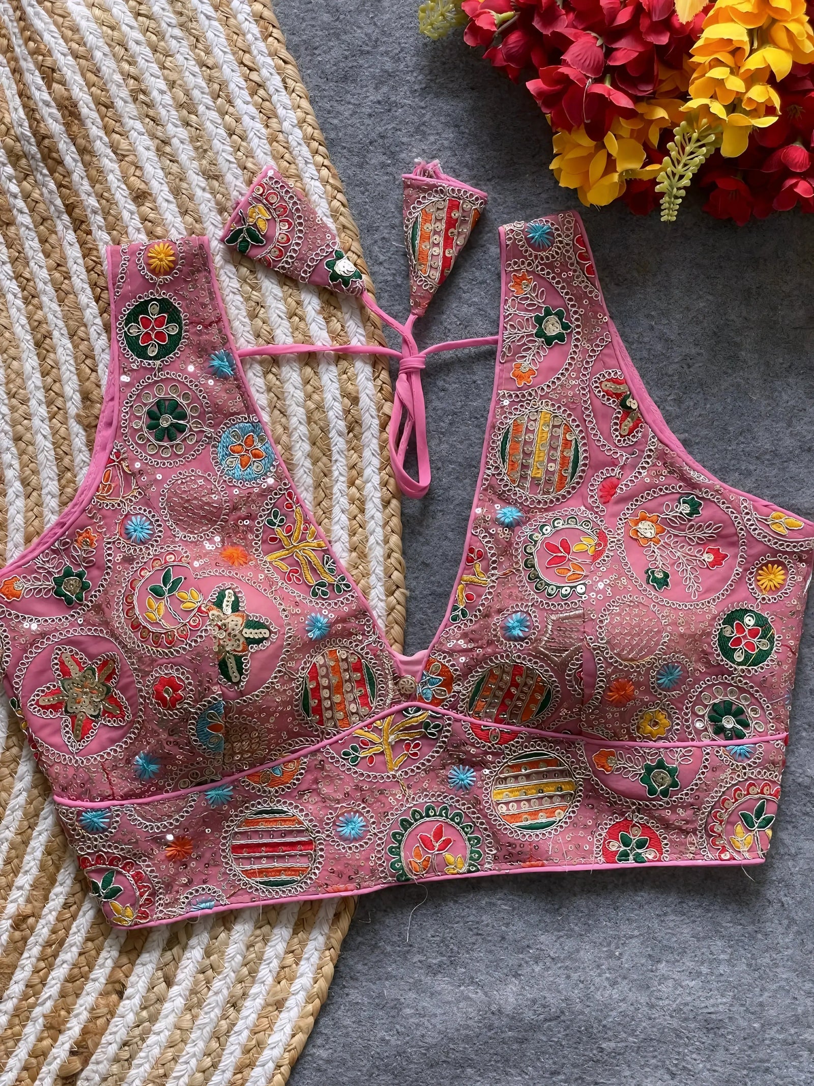Traditional pink blouse featuring vibrant multicolor thread embroidery and mirror work in circular floral and geometric patterns, with a deep V-neck and tie-back detail, displayed flat on a textured mat with artificial red and yellow flowers in the corner.