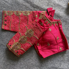 Traditional pink silk blouse featuring intricate gold embroidery and beadwork with floral and paisley motifs on the sleeves and neckline, displayed flat on a textured surface with a bunch of artificial flowers and a woven mat in the background.