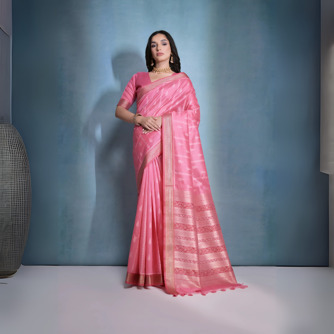 A woman models a striking Soft Pink Raw Silk Saree. The saree body features subtle woven motifs and is framed by a wide metallic gold Zari border. The pallu features intricate horizontal bands of pink and gold Ikat-style geometric weaving. She wears a matching short-sleeve pink blouse and accessorizes with a gold choker necklace and earrings, posing indoors.