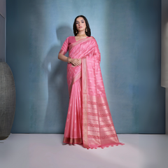 A woman models a striking Soft Pink Raw Silk Saree. The saree body features subtle woven motifs and is framed by a wide metallic gold Zari border. The pallu features intricate horizontal bands of pink and gold Ikat-style geometric weaving. She wears a matching short-sleeve pink blouse and accessorizes with a gold choker necklace and earrings, posing indoors.