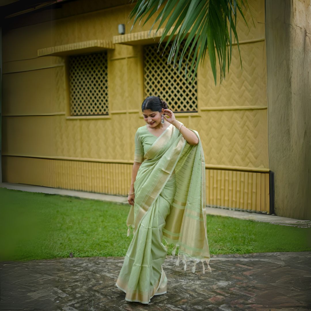 Woman in pista-colored zari work saree standing outdoors near a yellow building and palm leaves