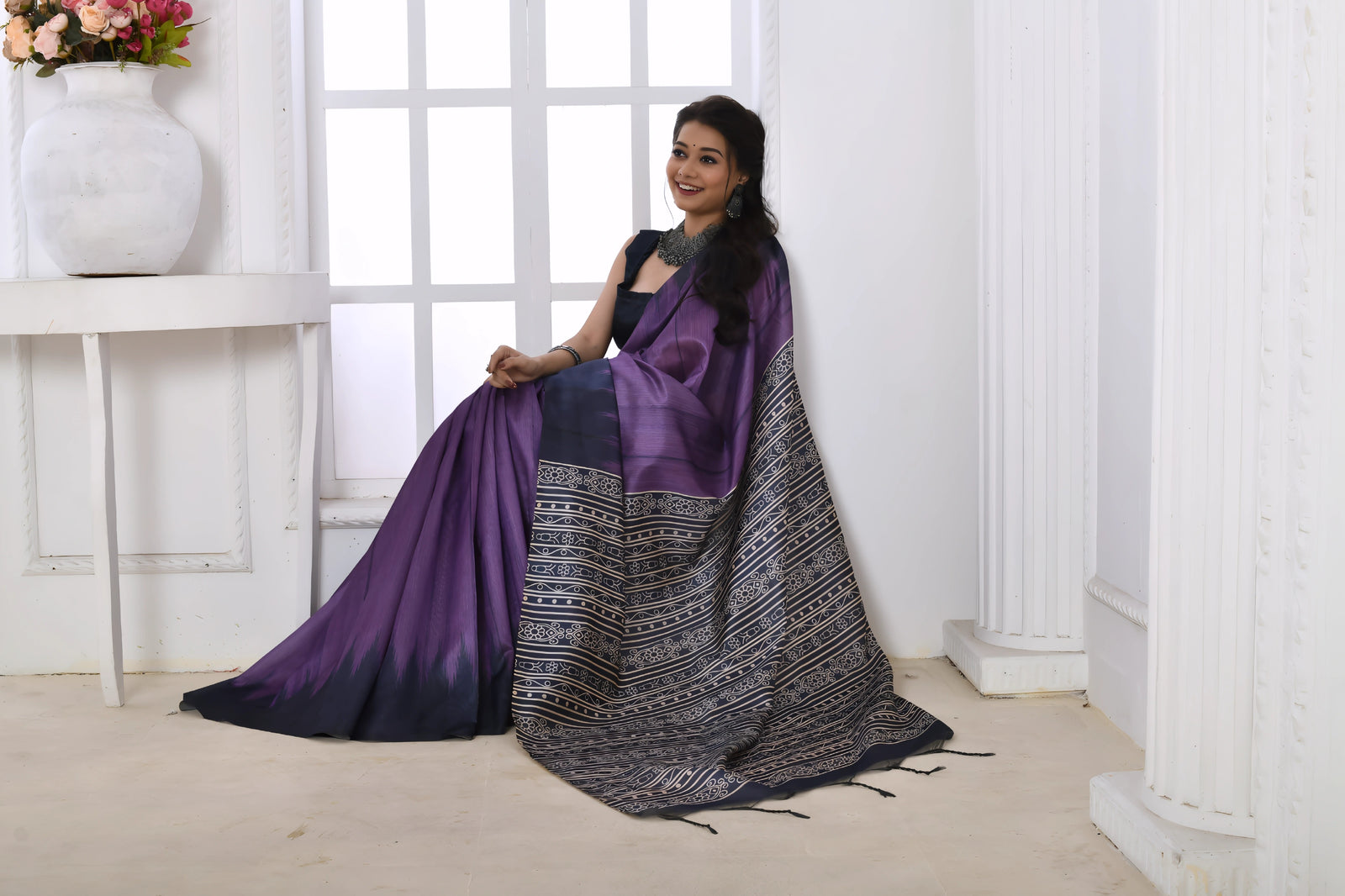A woman models a striking Purple Soft Tussar Silk Saree. The saree features a color-block transition between purple and deep navy/black. The pallu has a wide panel of dense, grey and black geometric Ikat-style print. She wears a dark sleeveless blouse and chunky oxidized silver jewelry.