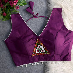 Purple padded blouse with white piping, featuring a central embroidered triangle motif with mirror work and shell detailing along the hem, displayed on a grey surface with artificial pink flowers and white fur in the background.
