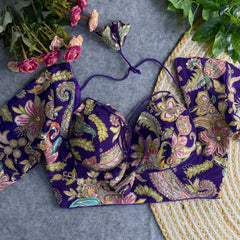 Purple padded blouse with multicolor floral and paisley embroidery, featuring a tie-back detail, displayed on a grey surface with artificial pink flowers and green leaves in the background.