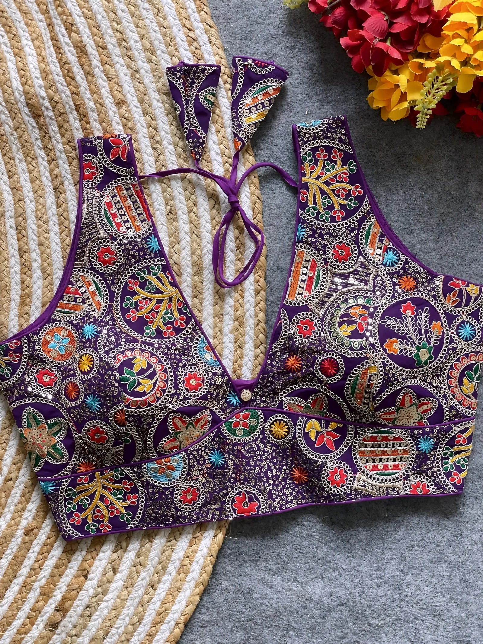 Traditional purple blouse featuring vibrant Madhubani-inspired multicolor embroidery with circular floral and geometric motifs, deep V-neck, and tie-back detail, displayed flat on a textured mat with artificial red and yellow flowers in the corner.