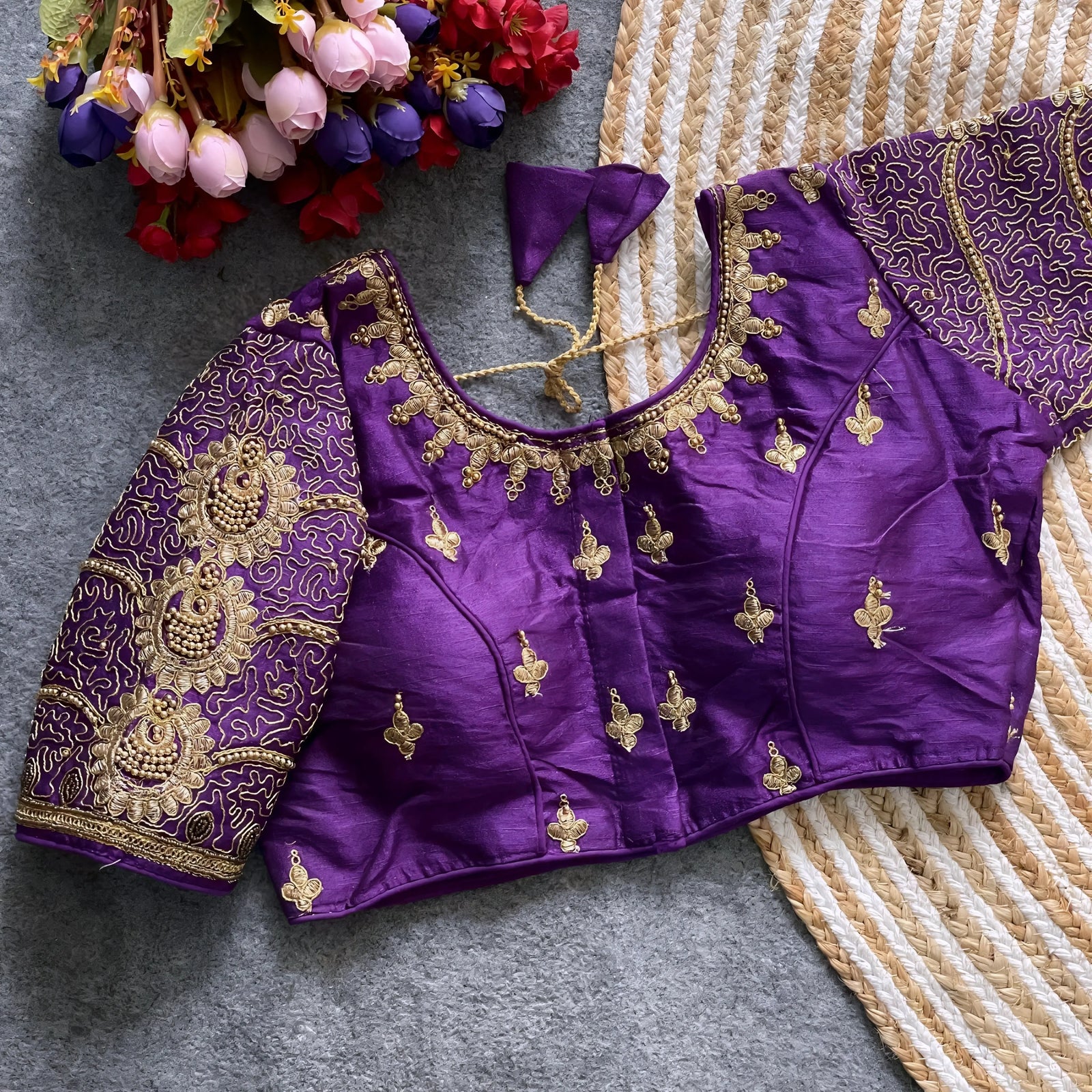 Traditional purple silk blouse featuring intricate gold embroidery and beadwork with floral and paisley motifs on the sleeves and neckline, displayed flat on a textured surface with a bunch of artificial flowers and a woven mat in the background.