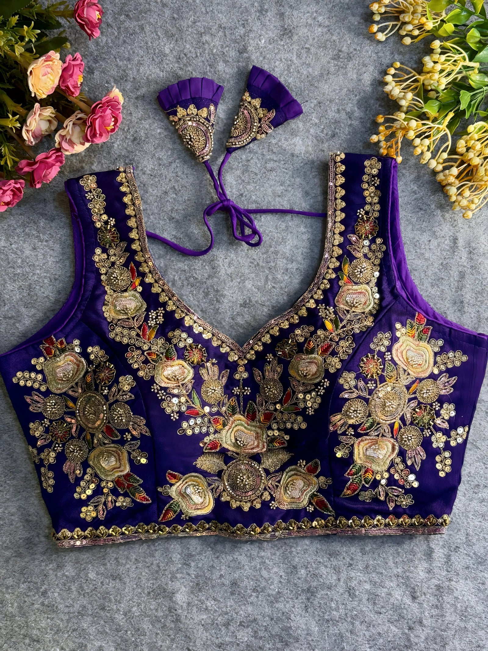 Traditional purple silk blouse featuring intricate gold zardozi embroidery and beadwork with floral and leaf motifs on the front and neckline, finished with decorative tassel ties, displayed flat on a textured grey background with artificial pink and yellow flowers in the corners.