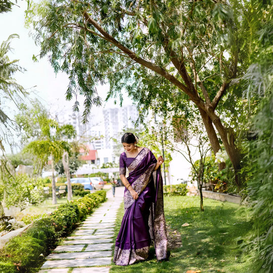 A woman models a luxurious Royal Purple Raw Silk Saree. The saree features a wide border and pallu with dense metallic (silver/light gold) Zari weaving in a Kalamkari-style floral and paisley pattern. She wears a matching purple short-sleeve blouse and gold jewelry, posing outdoors on a stone pathway.