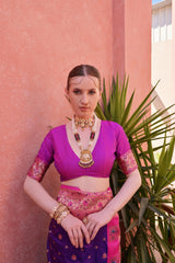 A woman models a vibrant Banarasi Silk Saree. The saree is royal purple with gold Zari weaving across the body, featuring a wide, ornate hot pink and gold Zari woven border and pallu. She pairs it with a short-sleeve pink blouse and heavy traditional gold jewelry.