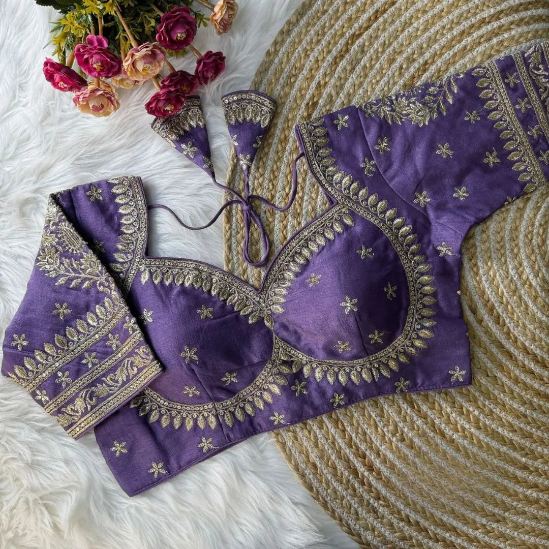 Purple blouse with intricate gold zari embroidery, featuring floral and leaf motifs, decorative sleeve borders, and tie-up tassels, displayed on a textured mat with pink flowers in the background.