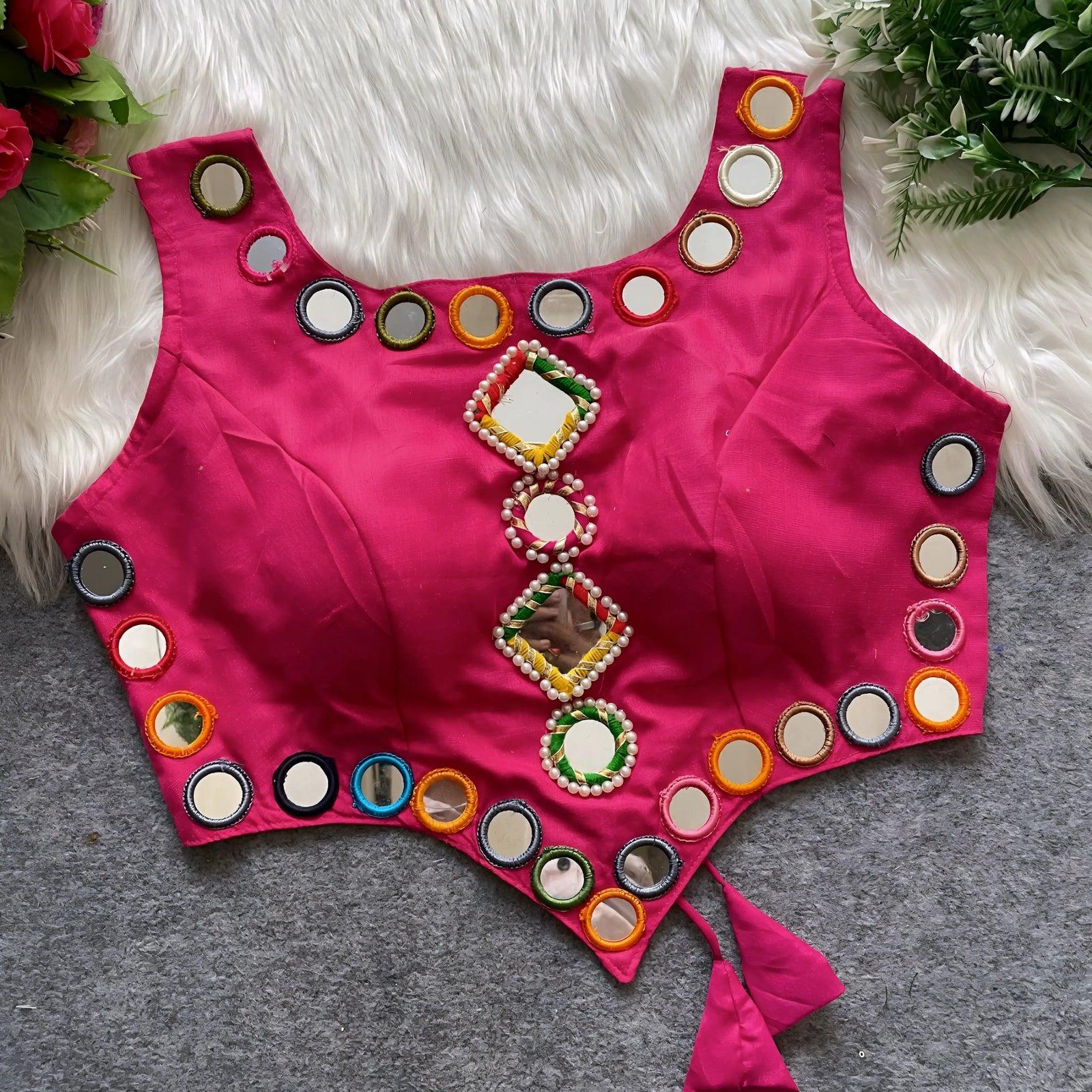Rani pink ethnic blouse featuring vibrant mirror work with multicolored embroidered borders and decorative diamond and round mirrors arranged vertically at the center, accented with pearl detailing and a tie-up tassel at the waist. Displayed flat on a grey surface with a white faux fur background and green leafy accents, highlighting its festive and traditional design.