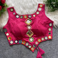Rani pink ethnic blouse featuring vibrant mirror work with multicolored embroidered borders and decorative diamond and round mirrors arranged vertically at the center, accented with pearl detailing and a tie-up tassel at the waist. Displayed flat on a grey surface with a white faux fur background and green leafy accents, highlighting its festive and traditional design.