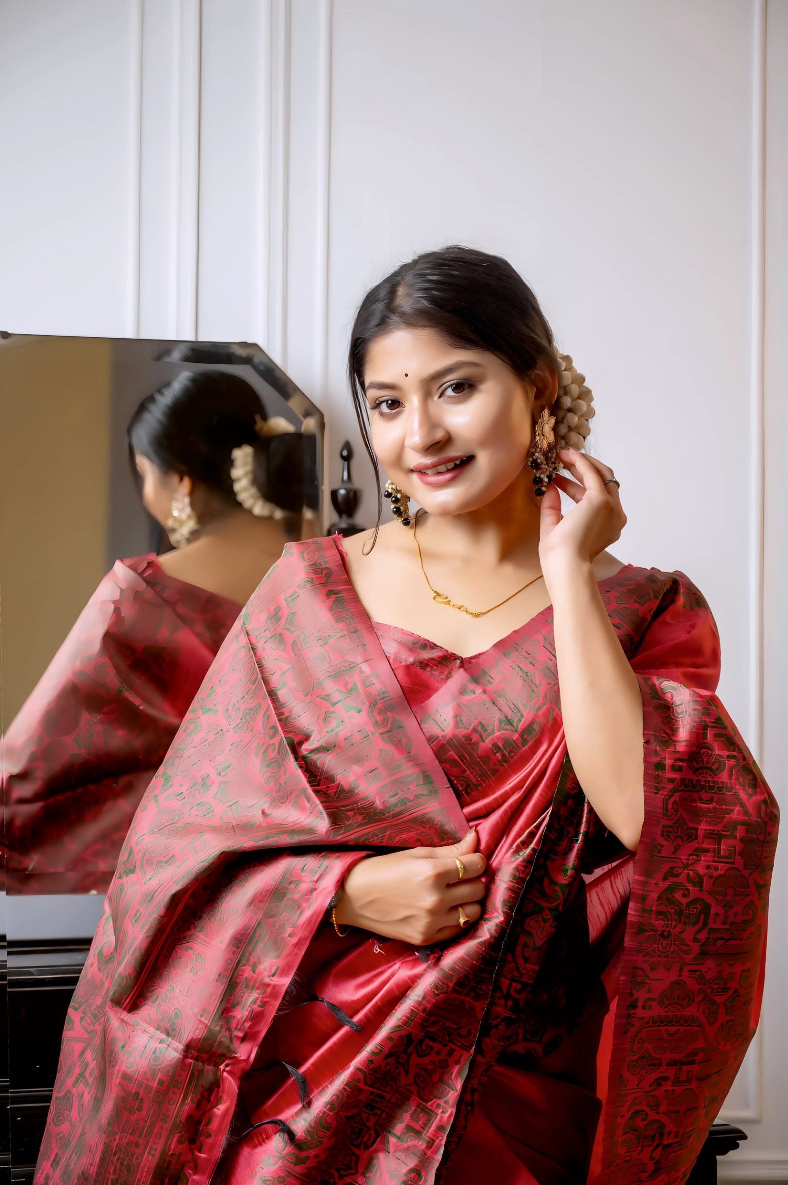 A woman models a luxurious Coral Red Raw Silk Saree. The saree features a wide border and pallu with dense, intricate weaving in a contrasting dark brown/black Kalamkari-style pattern. She wears a matching short-sleeve red blouse and traditional gold jewelry, with white flowers adorning her hair, posing indoors next to a dark wooden vanity.