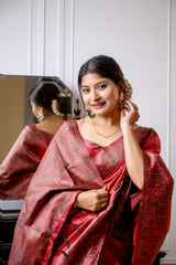 A woman models a luxurious Coral Red Raw Silk Saree. The saree features a wide border and pallu with dense, intricate weaving in a contrasting dark brown/black Kalamkari-style pattern. She wears a matching short-sleeve red blouse and traditional gold jewelry, with white flowers adorning her hair, posing indoors next to a dark wooden vanity.