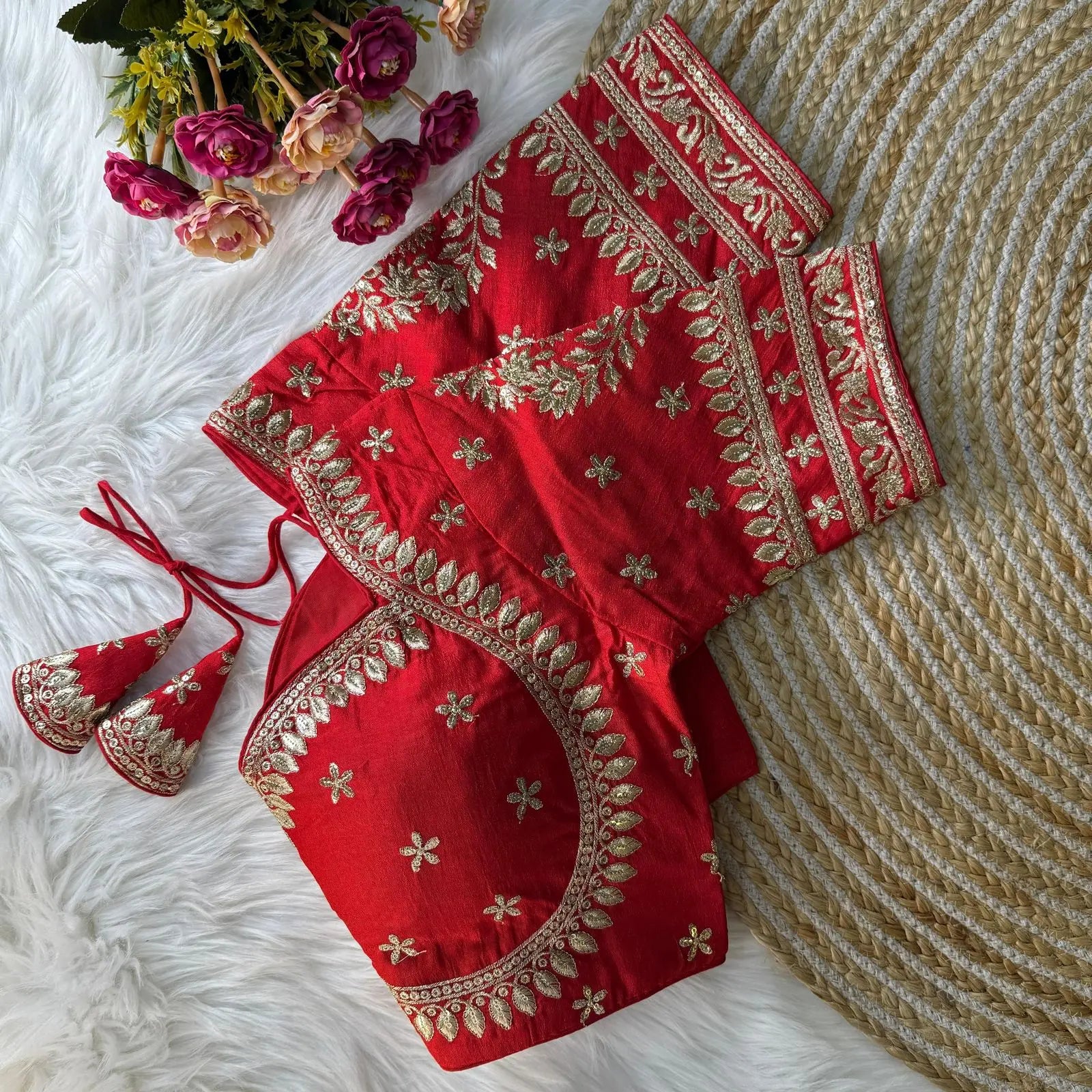 Red embroidered silk blouse with intricate gold zari floral patterns and tie-back detail, displayed on a white fur background with a bunch of pink and peach flowers in the corner
