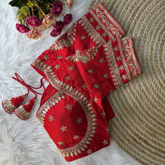 Red embroidered silk blouse with intricate gold zari floral patterns and tie-back detail, displayed on a white fur background with a bunch of pink and peach flowers in the corner