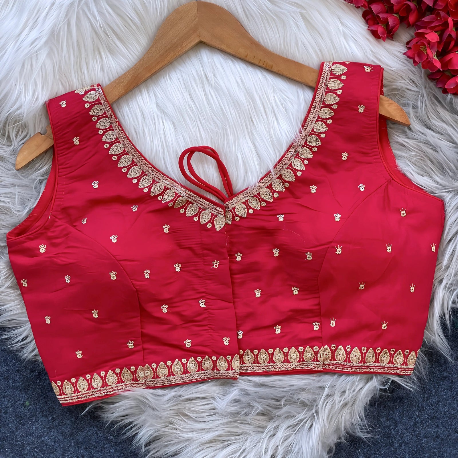 Red padded blouse with intricate gold embroidery along the neckline and hem, featuring small gold embroidered motifs, displayed on a wooden hanger over a white fur surface with red artificial flowers in the background.