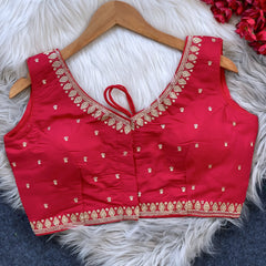 Red padded blouse with intricate gold embroidery along the neckline and hem, featuring small gold embroidered motifs, displayed on a wooden hanger over a white fur surface with red artificial flowers in the background.