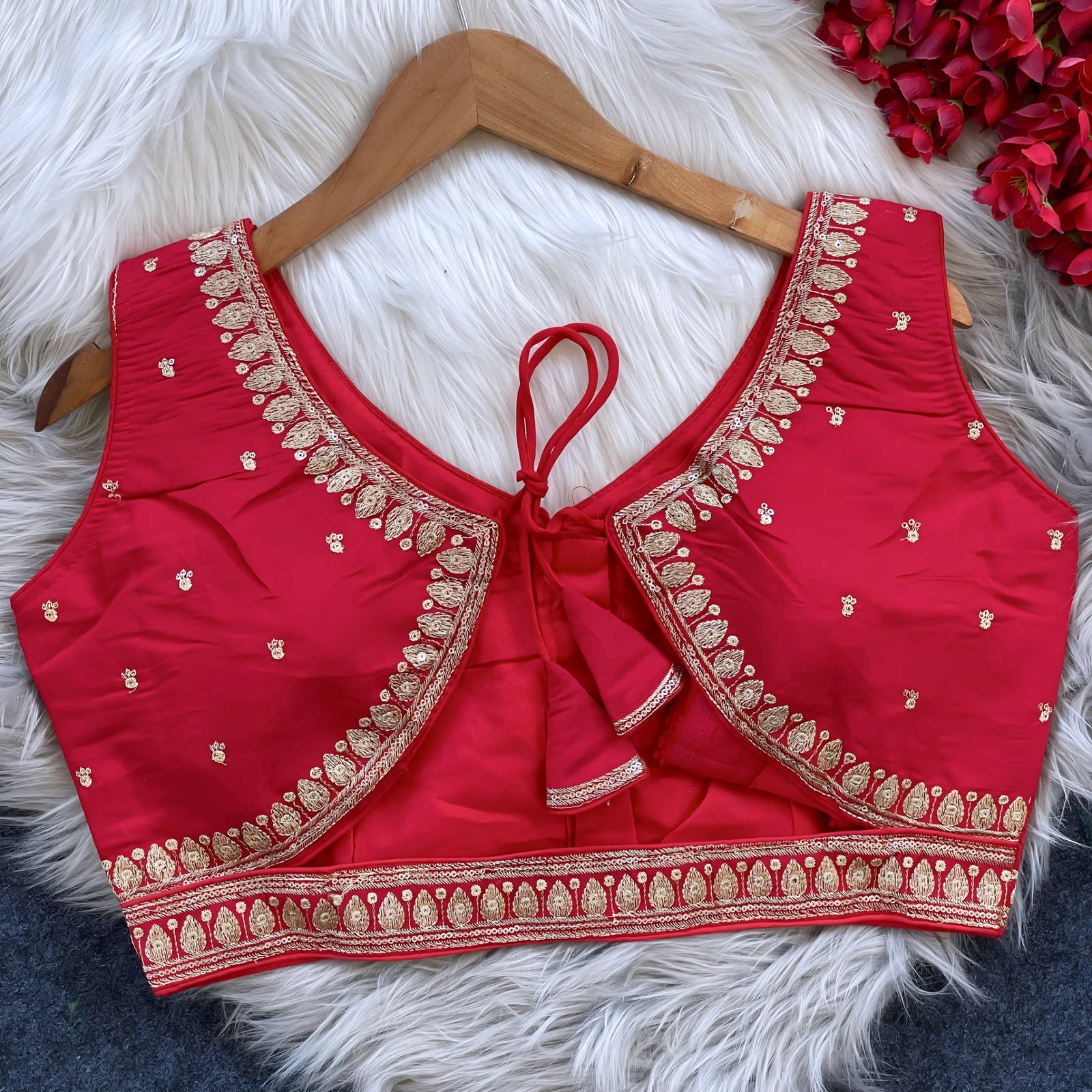 Red padded blouse with intricate gold embroidery along the neckline and hem, featuring small gold embroidered motifs, displayed on a wooden hanger over a white fur surface with red artificial flowers in the background.
