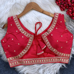 Red padded blouse with intricate gold embroidery along the neckline and hem, featuring small gold embroidered motifs, displayed on a wooden hanger over a white fur surface with red artificial flowers in the background.