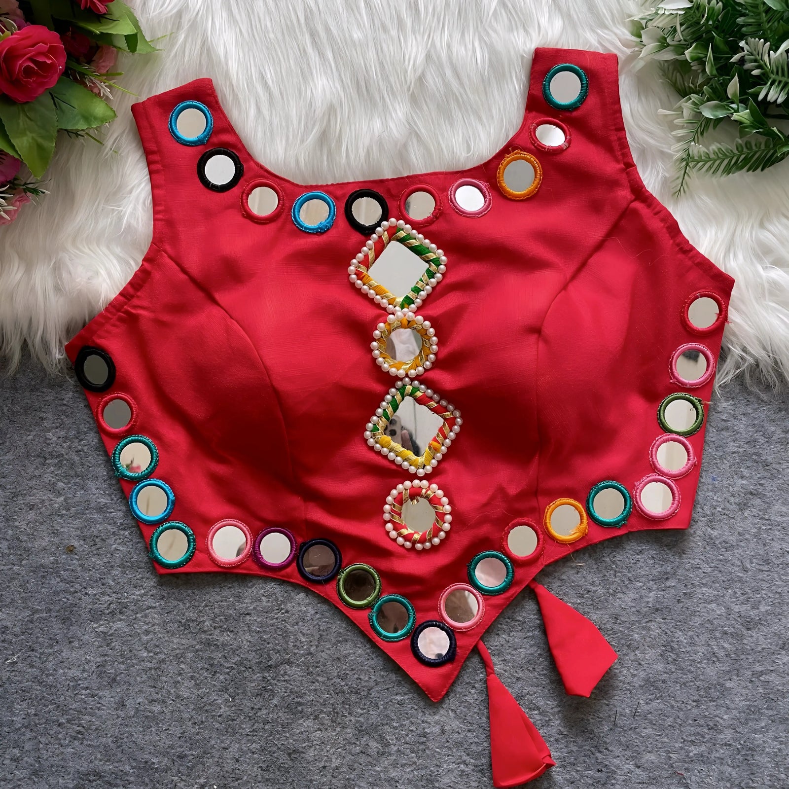 Red ethnic blouse featuring intricate mirror work with multicolored embroidered borders and embellished diamond-shaped mirrors at the center, finished with decorative tassel tie-ups at the waist. Displayed flat on a grey surface with white faux fur and floral accents for a festive and traditional look.