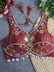 Traditional red blouse featuring vibrant multicolor mirror work, intricate thread embroidery in geometric and floral patterns, and shell tassel detailing along the hem, displayed on a flat surface with floral and leafy decor.