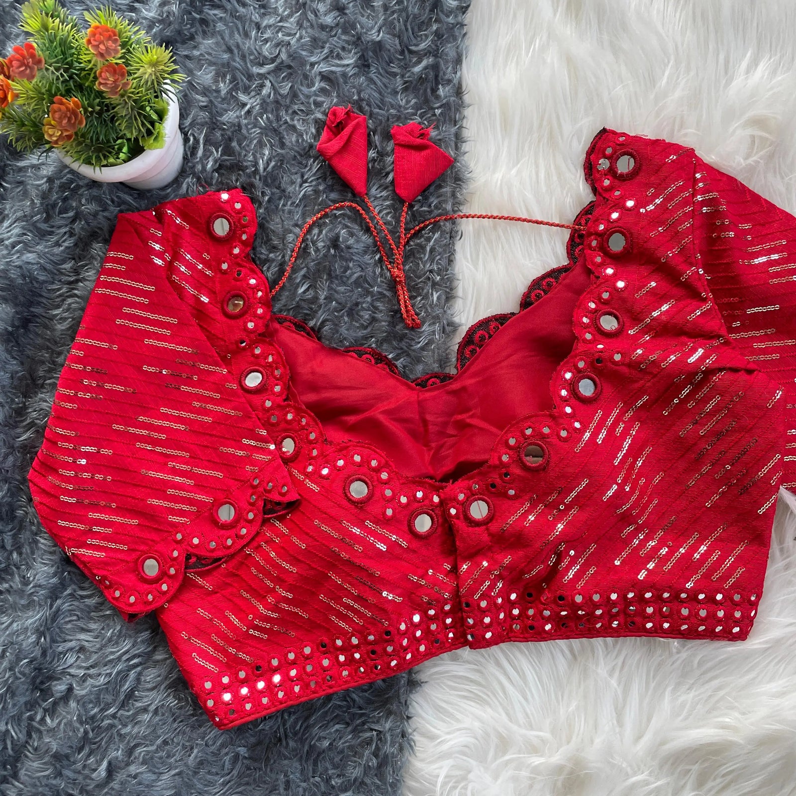 Red bridal blouse with scallop sleeves, featuring silver sequin stripes and prominent round mirror work along the neckline, sleeves, and hem, displayed on a textured grey and white fur background with a small potted plant nearby.