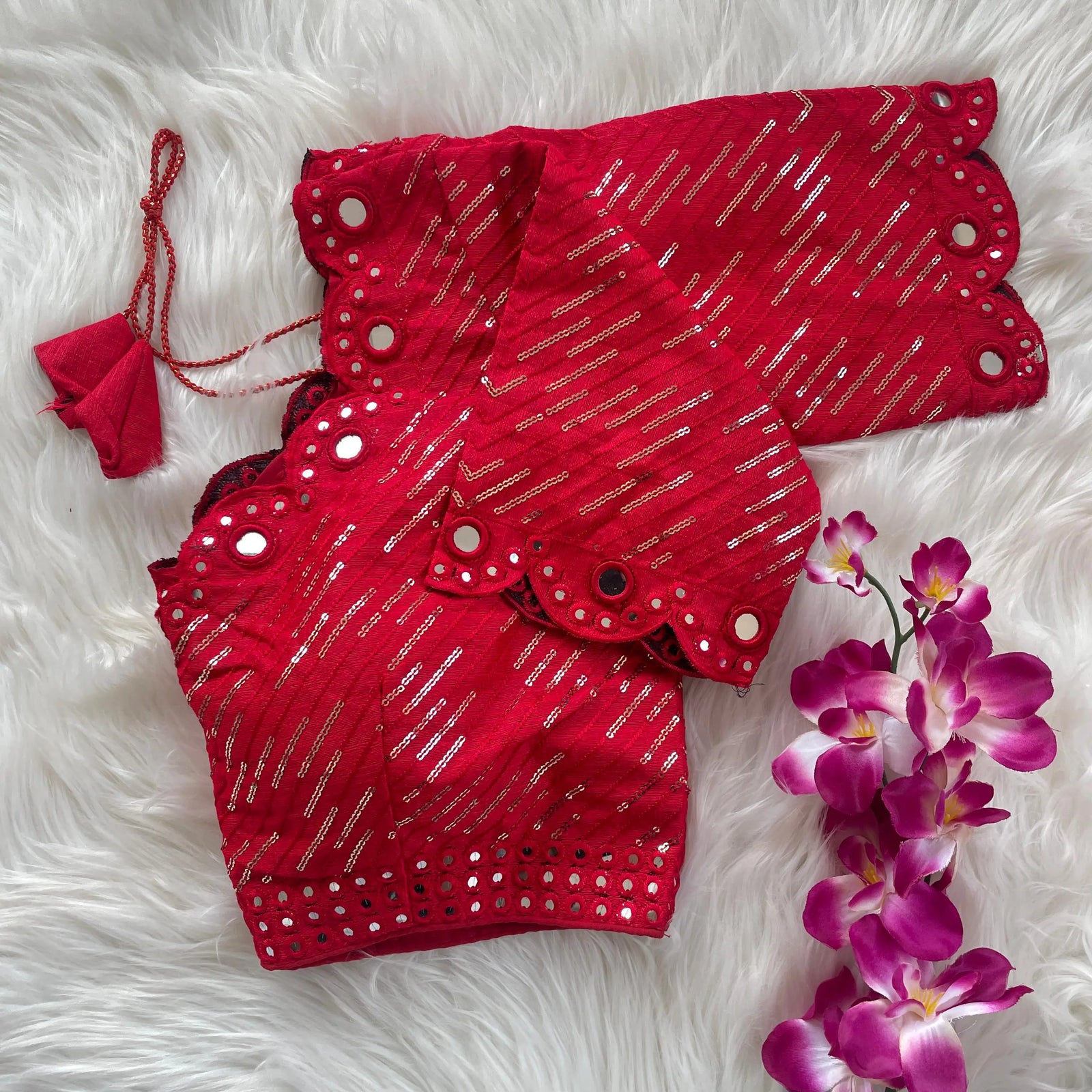 Red bridal blouse with scallop sleeves, featuring silver sequin stripes and prominent round mirror work along the neckline, sleeves, and hem, displayed on a textured grey and white fur background with a small potted plant nearby.