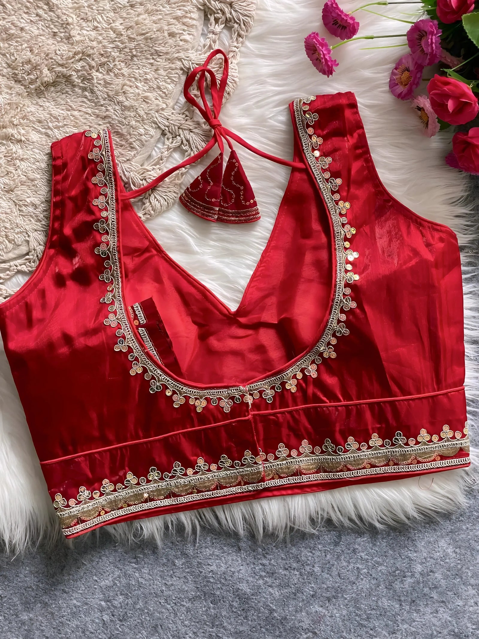 Traditional red silk blouse featuring intricate gold embroidery along the neckline and hem, with a decorative tassel tie at the back, displayed flat on a soft fur and textured mat background with artificial pink flowers in the corner.