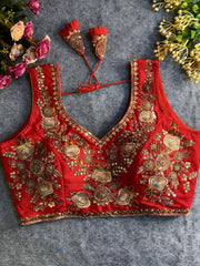Traditional red silk blouse featuring intricate gold zardozi embroidery and beadwork with multicolor floral and leaf motifs on the front and neckline, finished with decorative tassel ties, displayed flat on a textured grey background with artificial pink and yellow flowers in the corners.