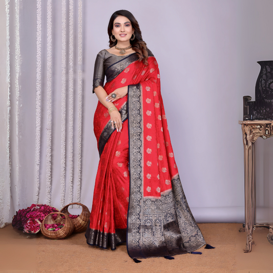 Woman wearing a striking red silk saree with intricate silver and black zari border, detailed buti motifs, and a matching black blouse. She is accessorized with traditional jewelry and bangles, posing elegantly in a contemporary studio setting with woven baskets of rose petals, sheer white curtains, and a neutral gray backdrop.