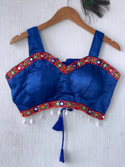 Royal blue ethnic blouse featuring vibrant mirror work and a multicolored embroidered geometric border in red, green, white, and orange, adorned with white pearl drop tassels and a tie-up waist detail. Displayed on a wooden hanger against a textured off-white wall with peacock feather, creating a festive and traditional look.