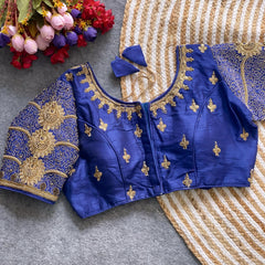 Traditional royal blue silk blouse featuring intricate gold embroidery and beadwork with floral and paisley motifs on the sleeves and neckline, displayed flat on a textured surface with a bunch of artificial flowers and a woven mat in the background.