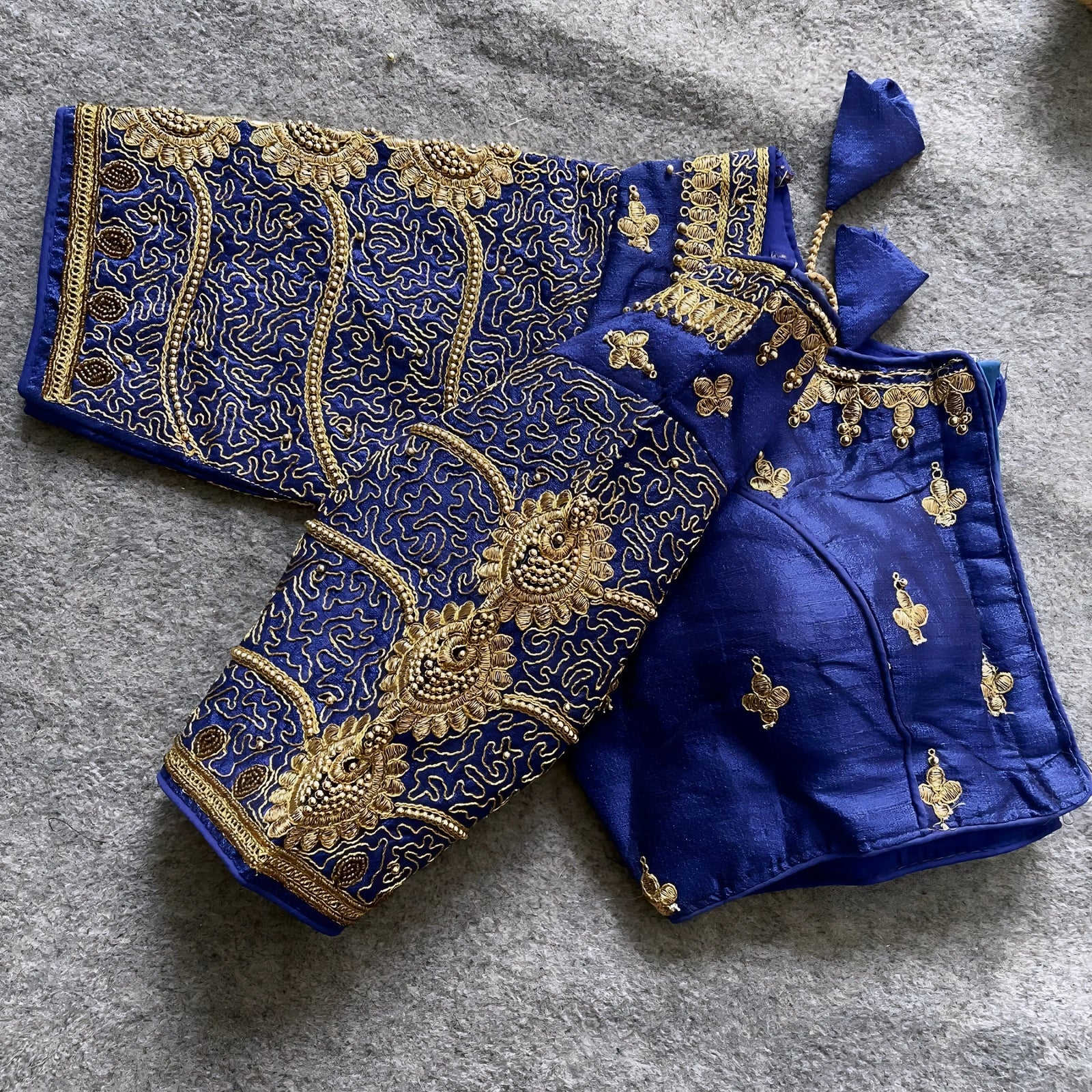 Traditional royal blue silk blouse featuring intricate gold embroidery and beadwork with floral and paisley motifs on the sleeves and neckline, displayed flat on a textured surface with a bunch of artificial flowers and a woven mat in the background.